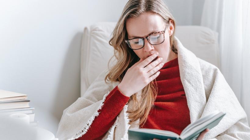 Eine junge Frau sitzt in einem Sessel, hat eine Decke über den Schultern, hält mit der einen Hand ein Buch, die andere Hand hält sie vor den Mund und gähnt.