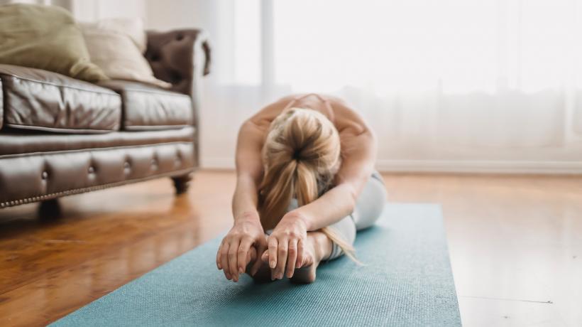 Vor einer Ledercouch liegt eine Yogamatte, auf der eine Frau eine Dehnungsübung macht.