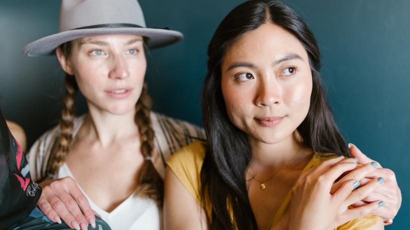 Zwei Frauen stehen entspannt nebeneinander, die eine hat der anderen die Hand auf die Schulter gelegt, die diese mit ihren eigenen Händen umfasst.