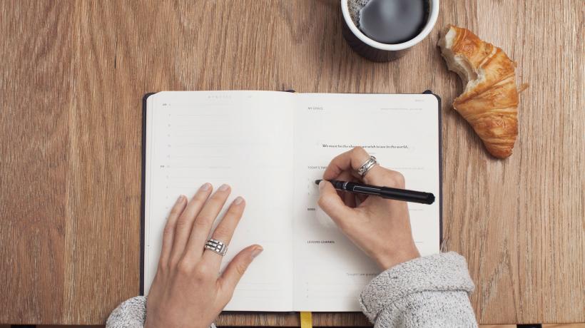 Blick von oben auf einen Schreibtisch mit Kaffee, Crossaints und Notizbuch, in das eine Person etwas schreibt.