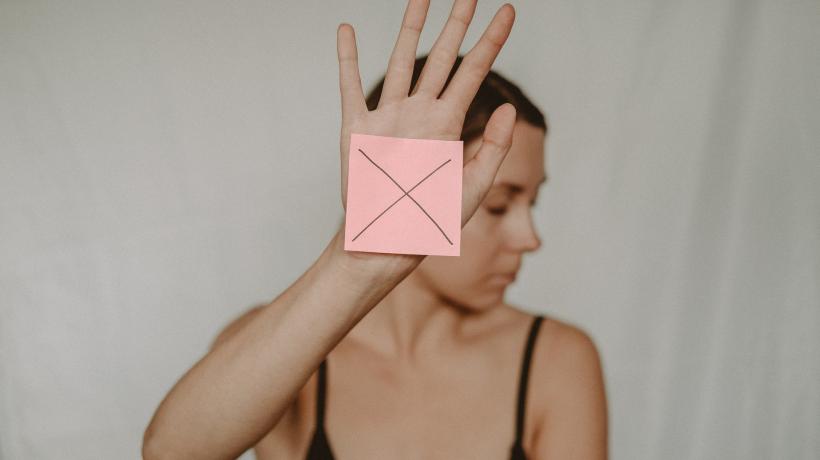 Eine Frau hält eine Hand vor sich, auf der ein Klebezettel mit einem großen X klebt, ihren Kopf wendet sie zur Seite weg.