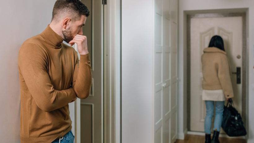 Eine Frau verlässt mit gepackter Tasche die Wohnung, etwas entfernt steht ein geknickter Mann im Flur und blickt ihr nach.
