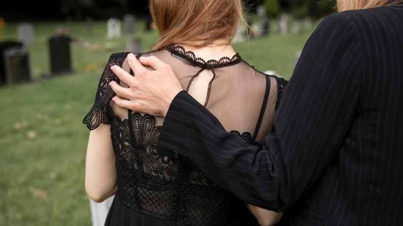 Zwei Personen in Trauerkleidung von hinten, die eine legt der anderen die Hand auf den Rücken.