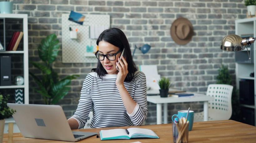Eine junge Frau sitzt in Ihrem Büro, telefoniert mit dem Handy und sitzt am Laptop